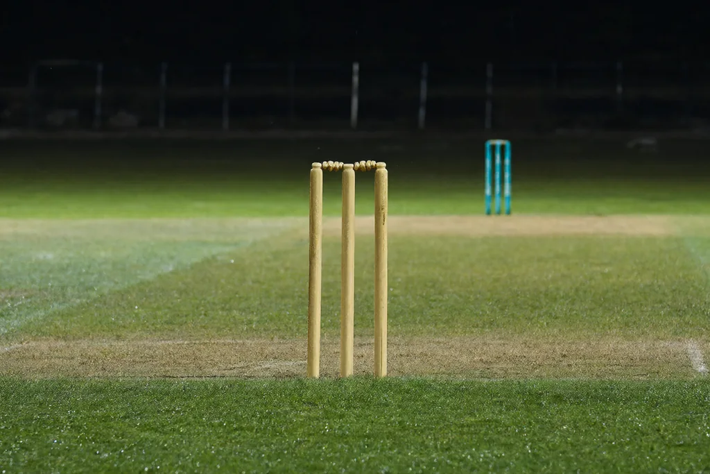 A cricket pitch under floodlights with dew on the grass
