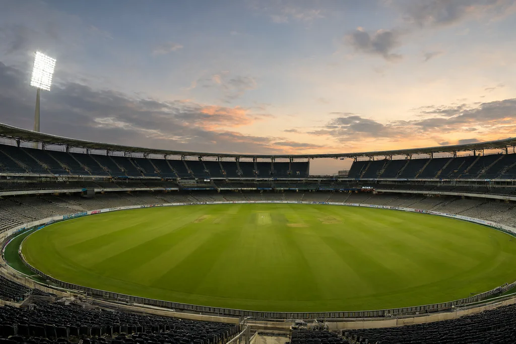 A cricket stadium at dusk with the floodlights warming up