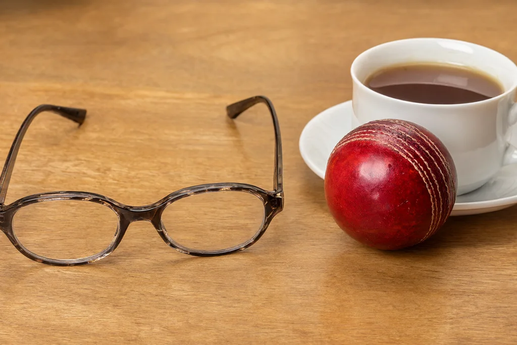 Reading glasses on a desk beside a cricket ball and a cup of tea
